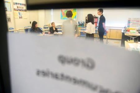 在前景是一扇门的玻璃窗户上一个模糊的纸标志, 在教室里，几个学生站起来对一组评委讲话.