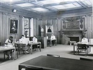 一个大房间的黑白照片，几张木桌排成一排. 有人坐在桌子旁看报纸. 