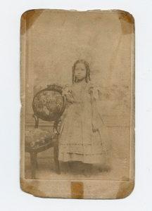 这是一张棕褐色的照片，一个年轻的女孩穿着长裙，手放在椅背上.