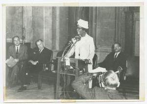 黑白照片，一个女人穿着白裙子，戴着白帽子在讲台上讲话. 两个男人坐在她的左边，一个坐在她的右边. 在左边的前景中，可以看到一个正在拍照的摄影师的背影.
