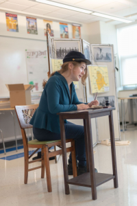 一个学生在教室里, 坐在一张小桌子前的椅子上，手里拿着一支笔, 身体前倾, 表演 
