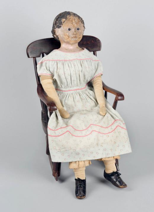 一个洋娃娃坐在摇椅上的照片. 娃娃穿着裙子和鞋子. 她的脸有裂缝，头发也涂上了脂粉.