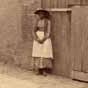 <p>Sepia photograph of a street and the side of a tall, 砖房，有四扇拱门和一盏大铁灯. 在大楼的左边, 披着披肩的黑人妇女, 帽子, 围裙, 她的长袖连衣裙靠墙站着，双手紧握在胸前.</p>